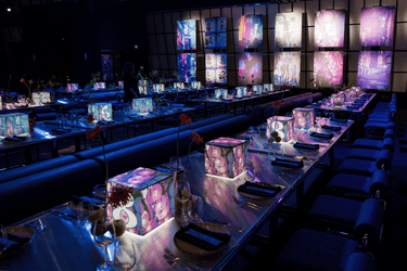 Dimly lit event space with long banquet tables set for dinner, featuring glowing cube centrepieces designed by Adelphi Mou with custom illustrated graphics and bold numbers. Each table is styled with modern tableware, single-stem flowers, and moody neon lighting. Large vertical panels with cityscape visuals line the back wall, enhancing the immersive atmosphere. Stationery and decor design by Adelphi Mou.